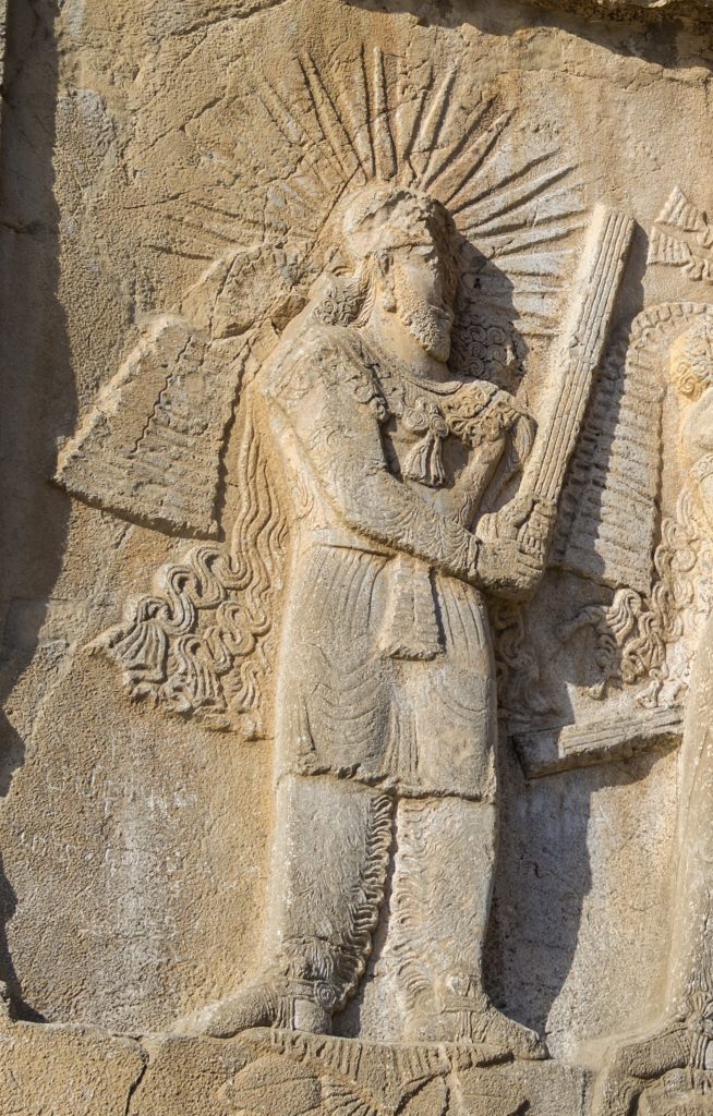 Mithras, dressed in Persian clothing, stands watching the investiture of Ardashir II (not pictured).  A halo is visible around his head.  He holds what appears to be a sword.  This is Mithras as the Persians imagined him.
